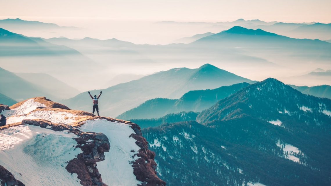 A hiker stands on a snowy mountain peak, arms raised in triumph, surrounded by a vast mountain range.