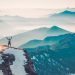 A hiker stands on a snowy mountain peak, arms raised in triumph, surrounded by a vast mountain range.